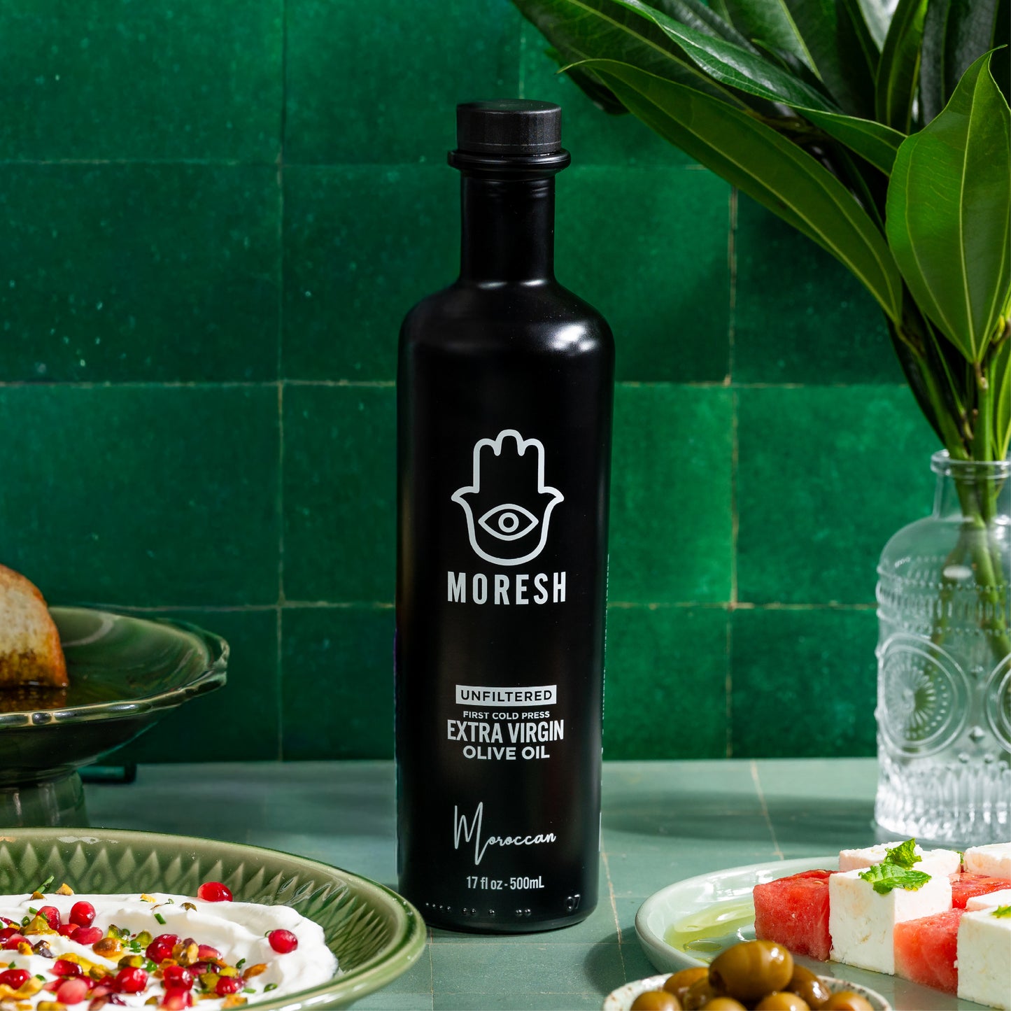 A bottle of MORESH unfiltered extra virgin olive oil stands on a countertop in a kitchen setting with green tile backsplash, surrounded by food props like olives, cheese, and a glass vase with leaves.