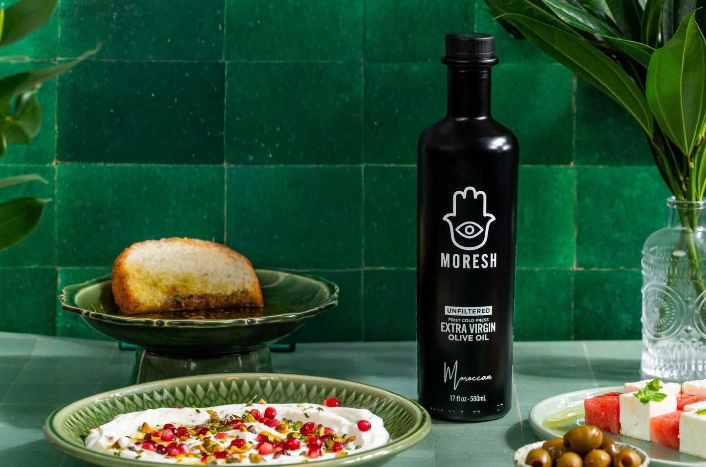 A bottle of Moresh Unfiltered Extra Virgin Olive Oil stands on a countertop in a styled kitchen scene with green tiles, a plate of dip topped with pomegranate seeds, bread, olives, and cheese, and a vase of leaves in the background.